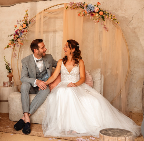 real bride sanneke – romantische A-lijn trouwjurk – bruid zit met haar partner op een sfeervol ingerichte zithoek; ronde backdrop met pastelbloemen vormt een dromerige achtergrond
